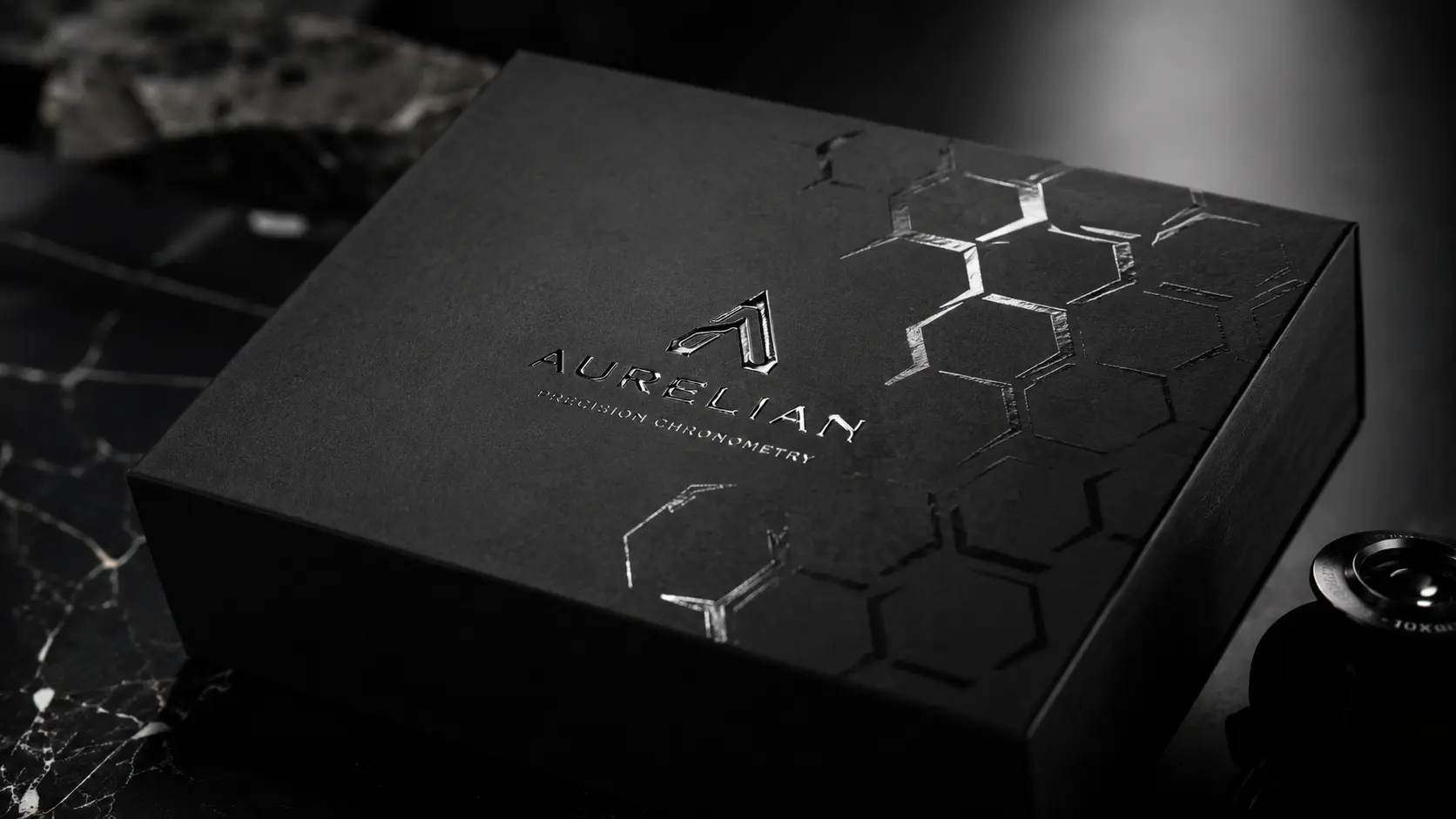 Caja negra premium de la marca Aurelia Precision Chronometry sobre una superficie oscura, con logotipo metálico, textura mate y patrones hexagonales en relieve que transmiten lujo y precisión. Barniz UV y Acabados Premium